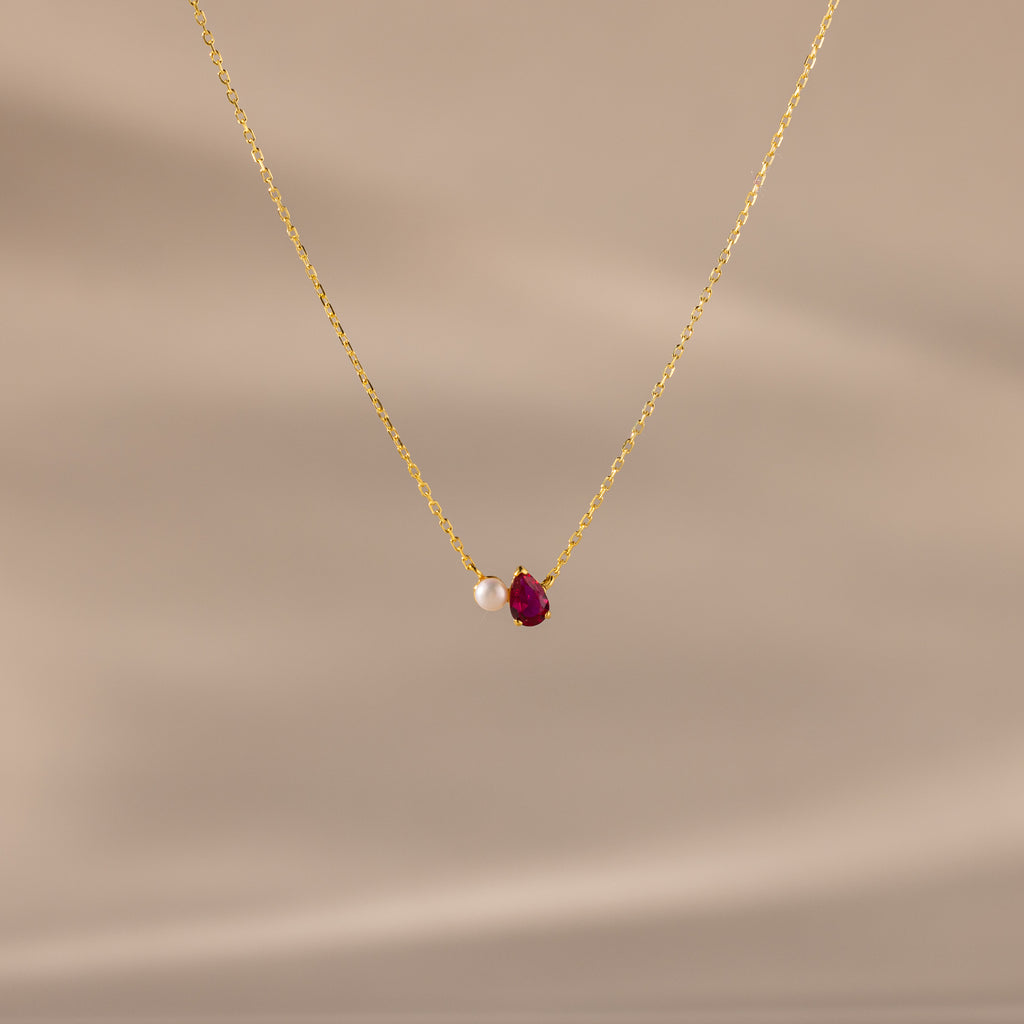The Teardrop Ruby & Pearl Necklace showcases a delicate pearl and red teardrop birthstone pendant on a soft beige backdrop—perfect for celebrating milestones.
