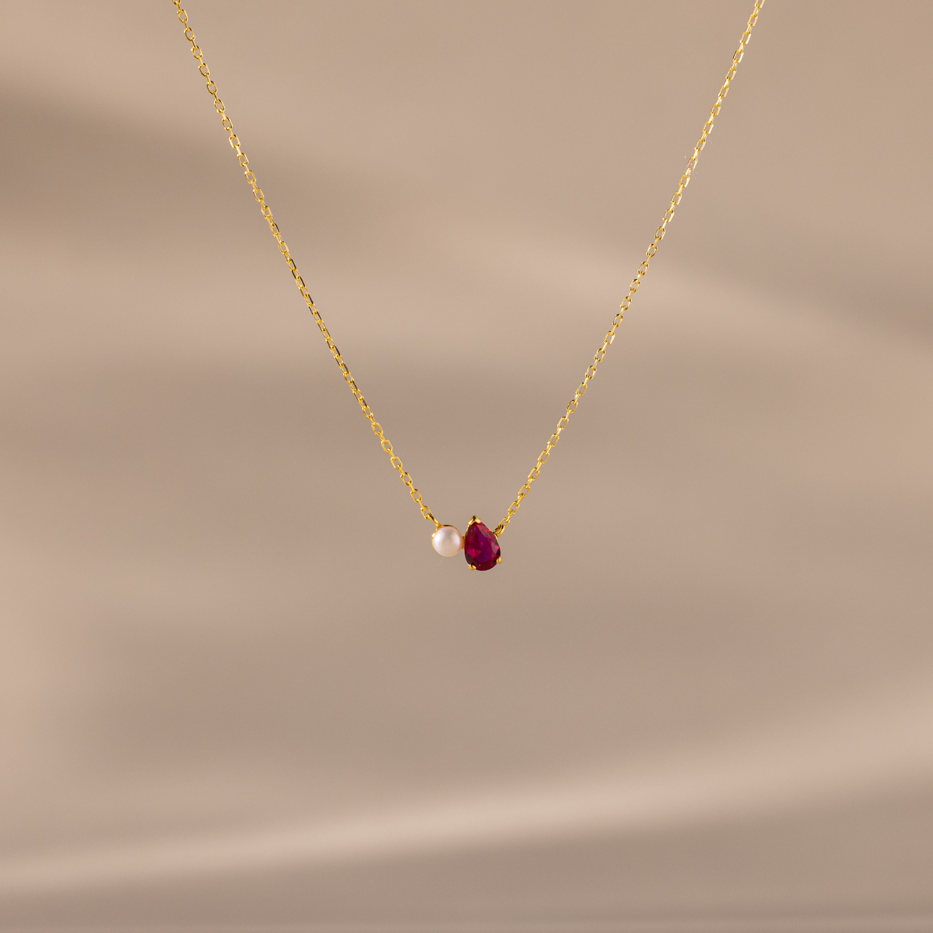 The Teardrop Ruby & Pearl Necklace showcases a delicate pearl and red teardrop birthstone pendant on a soft beige backdrop—perfect for celebrating milestones.