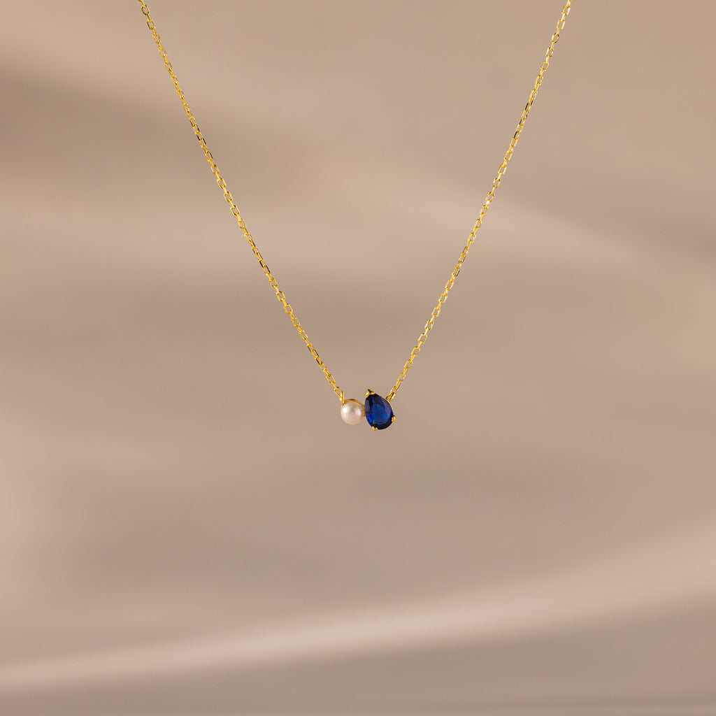 Teardrop Sapphire & Pearl Necklace with a gold chain, featuring a blue sapphire and a delicate pearl on a soft beige background—a timeless gift for any occasion.