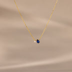 Teardrop Sapphire & Pearl Necklace with a gold chain, featuring a blue sapphire and a delicate pearl on a soft beige background—a timeless gift for any occasion.