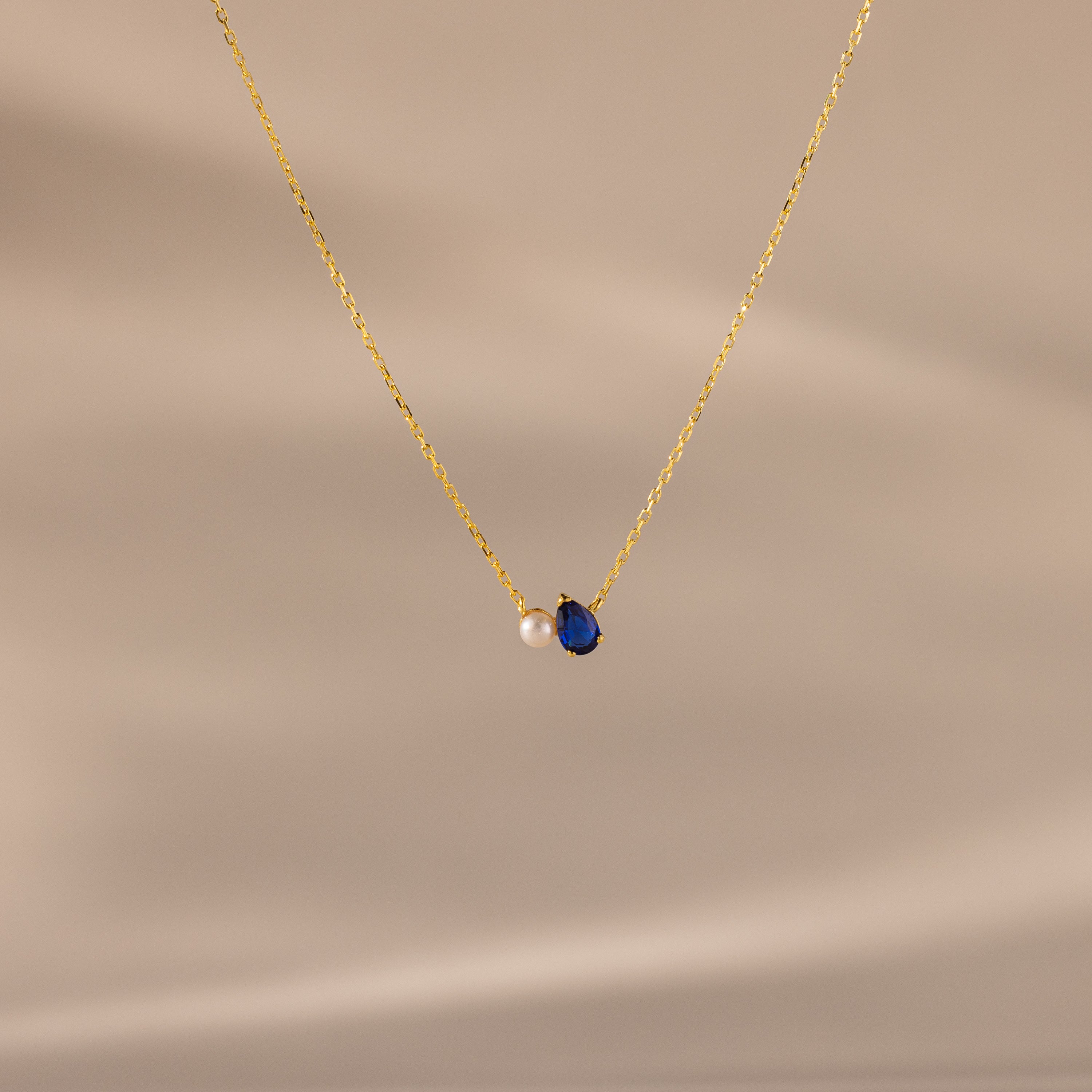 Teardrop Sapphire & Pearl Necklace with a gold chain, featuring a blue sapphire and a delicate pearl on a soft beige background—a timeless gift for any occasion.