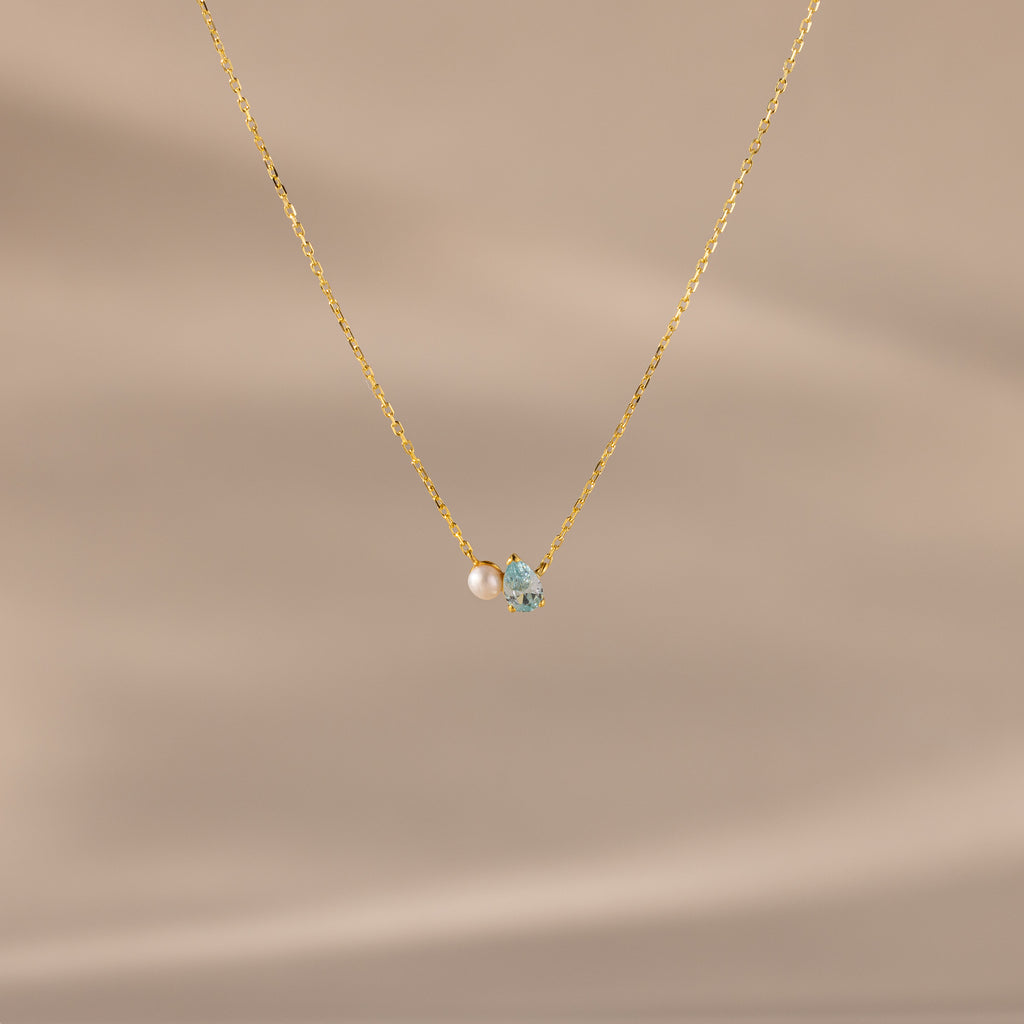 The Teardrop Aquamarine & Pearl Necklace features a small pearl and light blue aquamarine pendant, making it a perfect gift for milestones. Presented against a beige background.