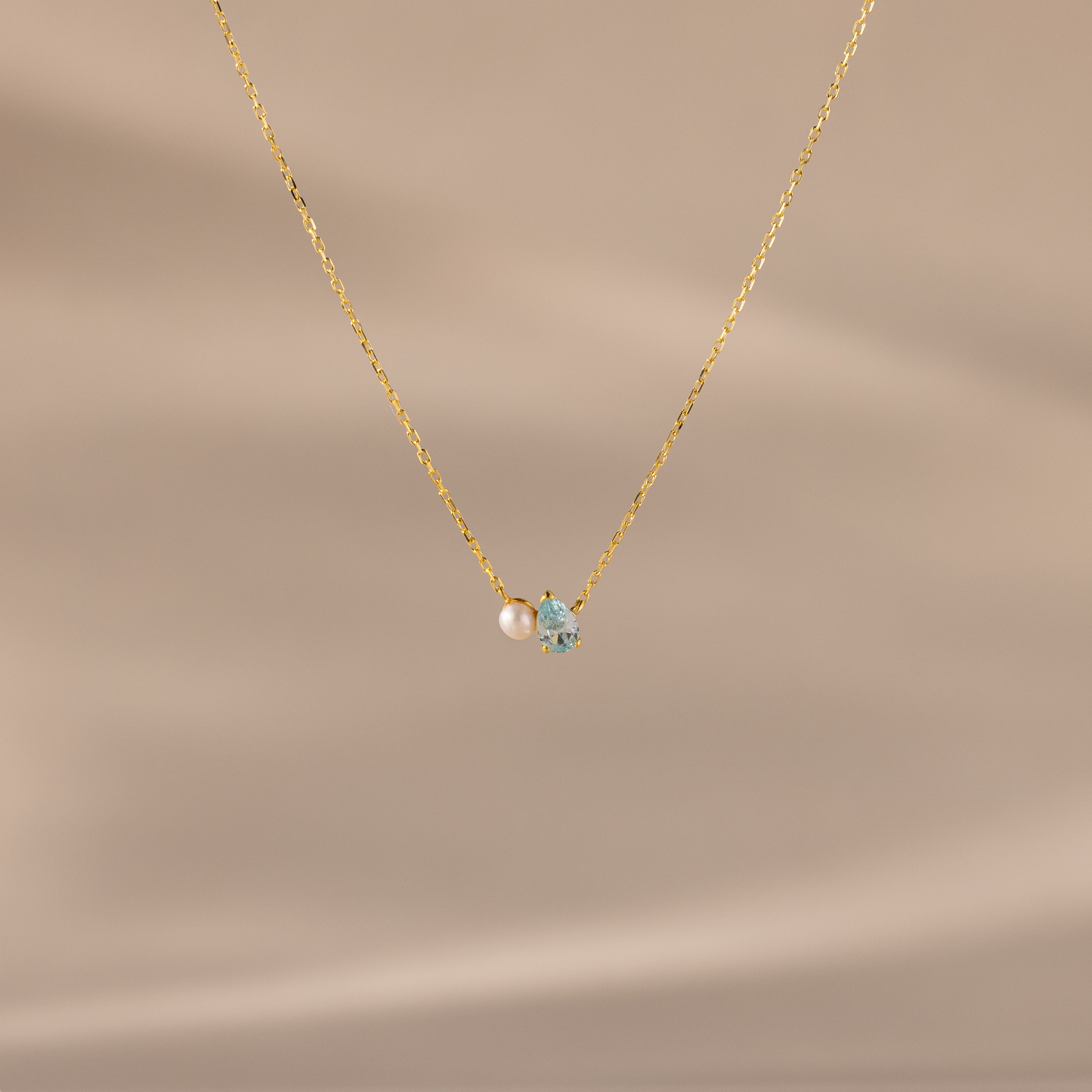 The Teardrop Aquamarine & Pearl Necklace features a small pearl and light blue aquamarine pendant, making it a perfect gift for milestones. Presented against a beige background.