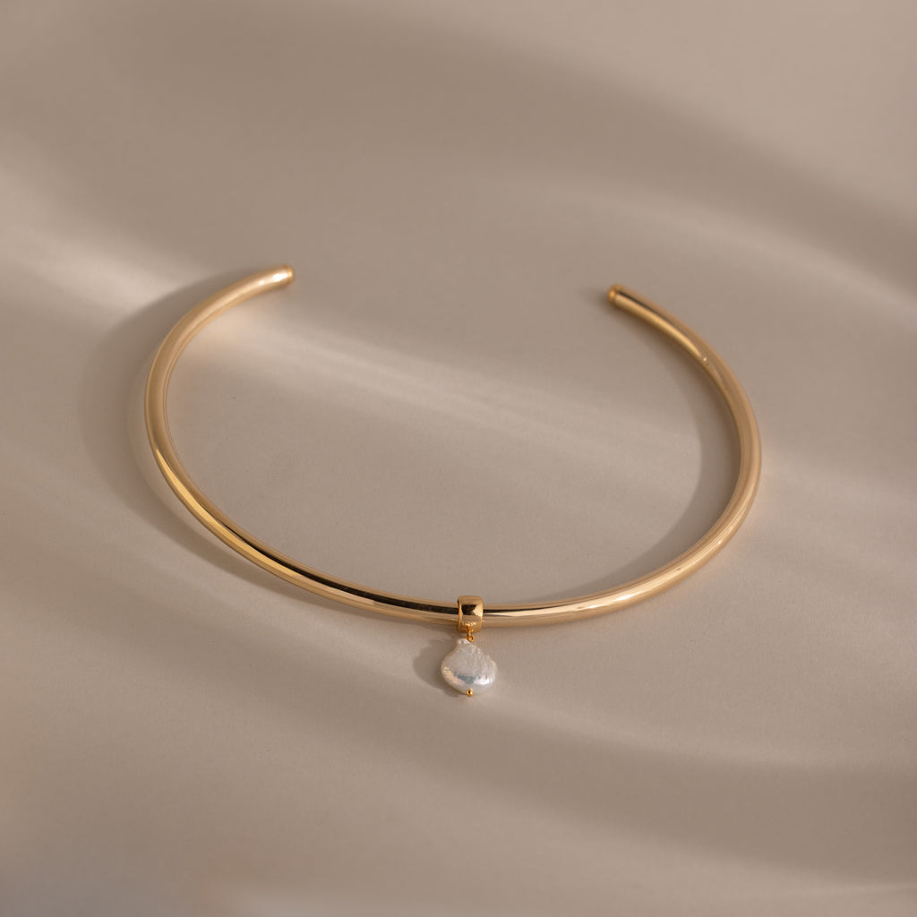 The Pearl Cuff Choker Necklace in 18K Gold features a small baroque pearl pendant, shown on a beige background with soft lighting.
