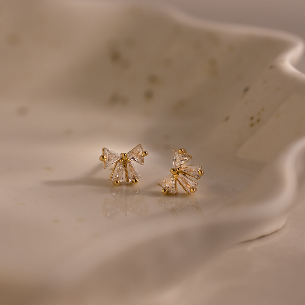 Close-up of gold bow stud earrings with clear crystal details, displayed on a ceramic dish.