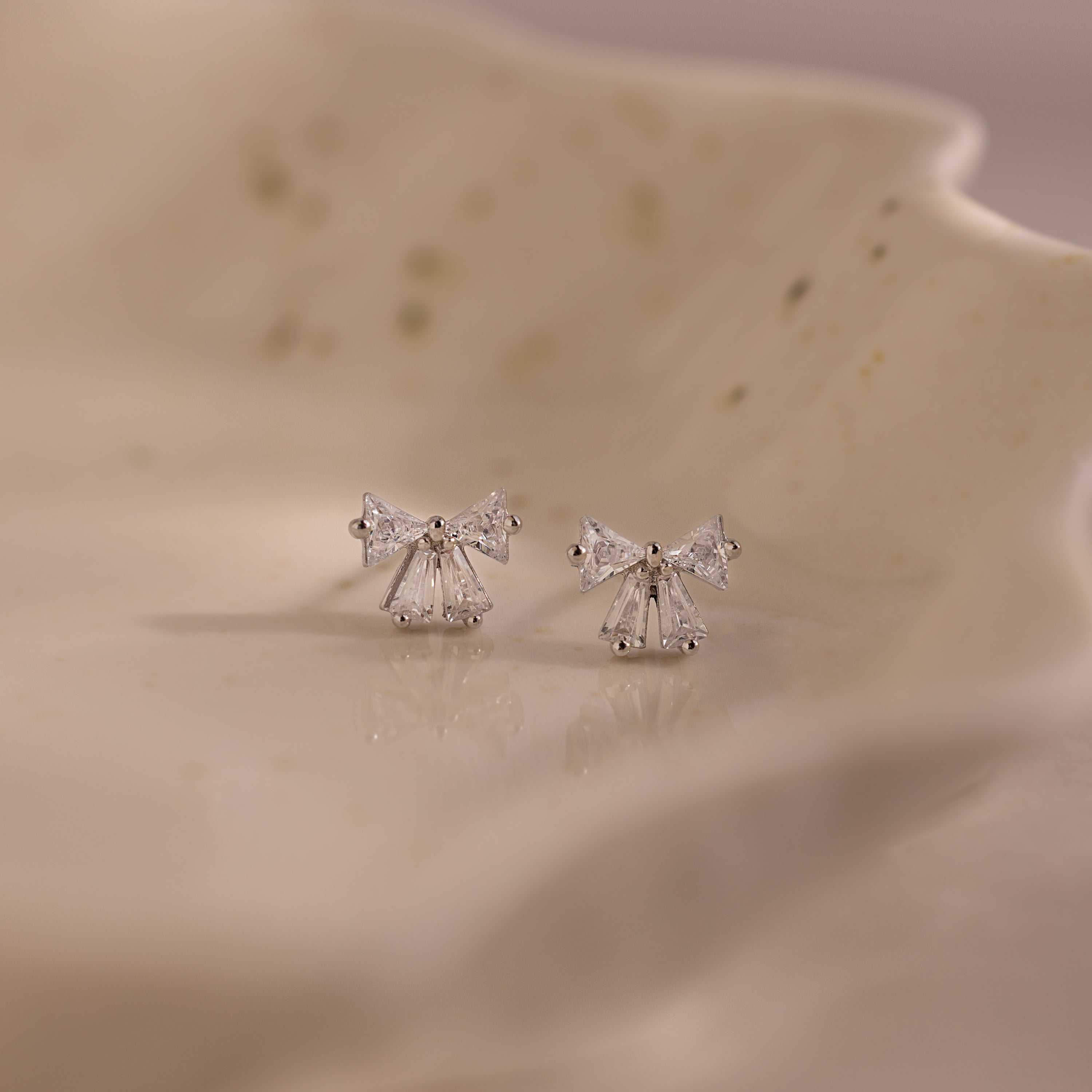 Close-up of silver bow stud earrings with clear crystal accents, displayed on a ceramic dish.