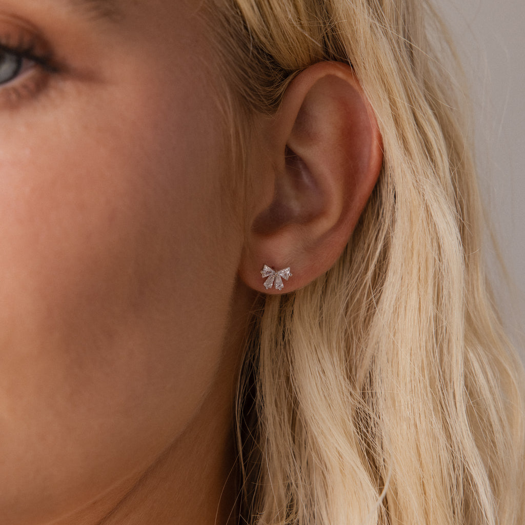 Close up of a woman wearing the Starlight Bow stud earring in silver, featuring baguette cut clear stones arranged in a delicate bow design for an elegant look.