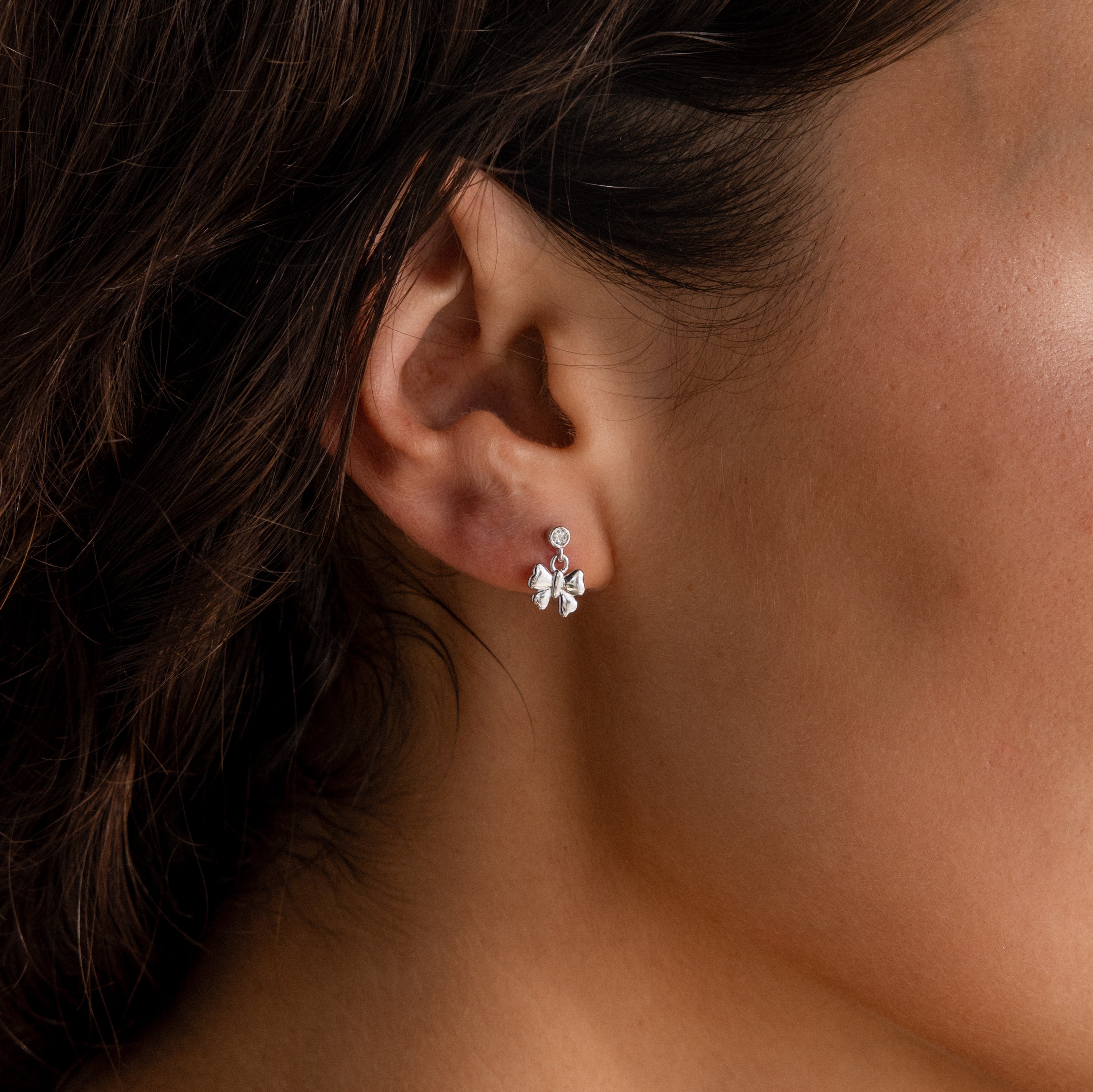 Close-up of a womans ear wearing the Bow Drop Studs in Sterling Silver, featuring a delicate flower-shaped design with a small round gemstone—perfect for fans of elegant styles.