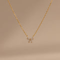 The Diamond Bow Necklace features a delicate gold-plated chain with a symmetrical bow pendant set with sparkling cubic zirconia, displayed on a soft beige background.