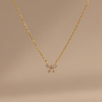 The Diamond Bow Necklace features a delicate gold-plated chain with a symmetrical bow pendant set with sparkling cubic zirconia, displayed on a soft beige background.