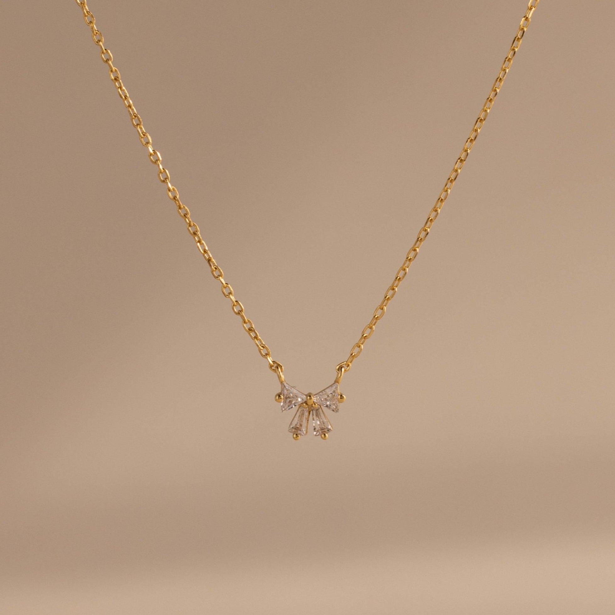 The Diamond Bow Necklace features a delicate gold-plated chain with a symmetrical bow pendant set with sparkling cubic zirconia, displayed on a soft beige background.