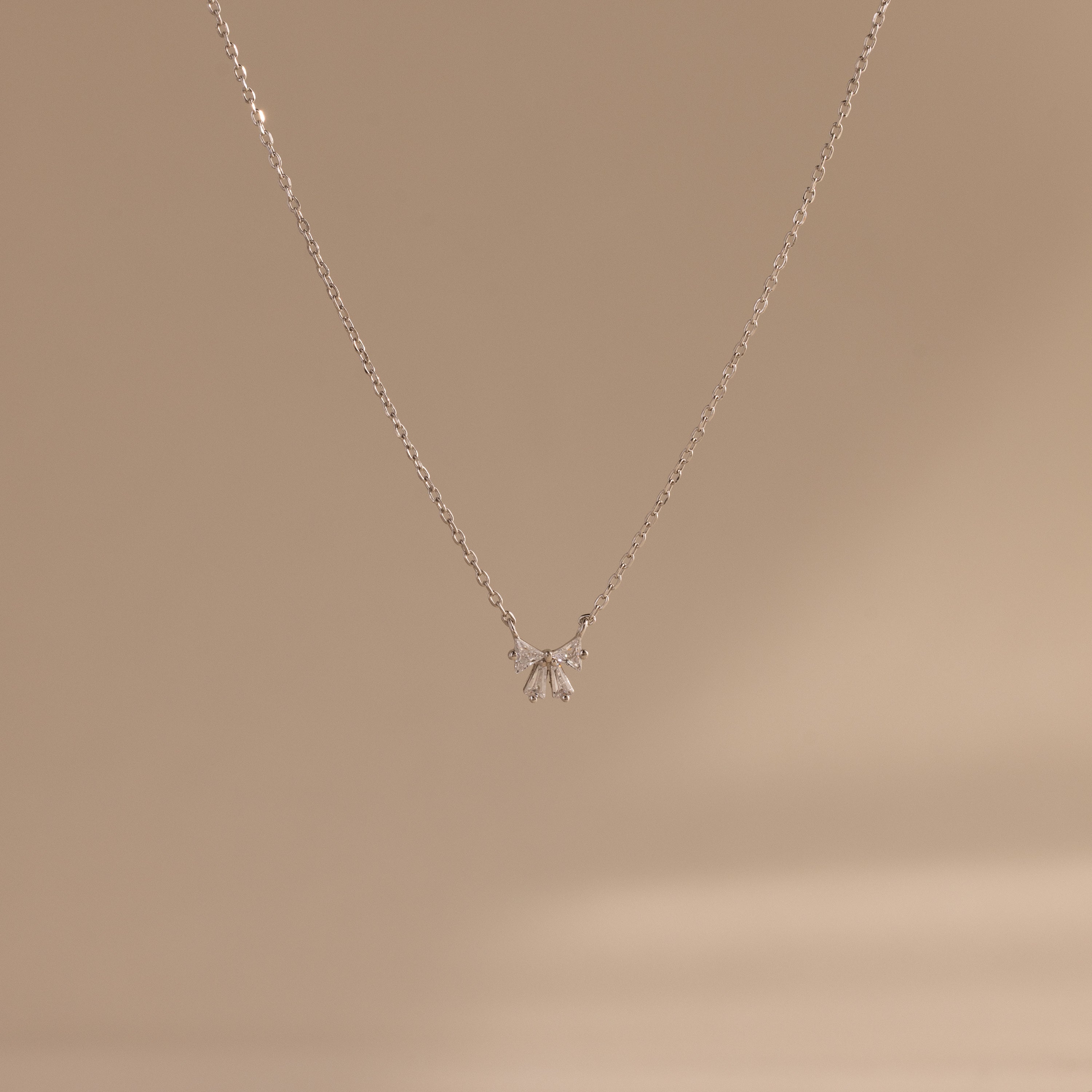 The Diamond Bow Necklace features a delicate gold-plated chain with a minimalist bow pendant, beautifully displayed on a soft beige background.