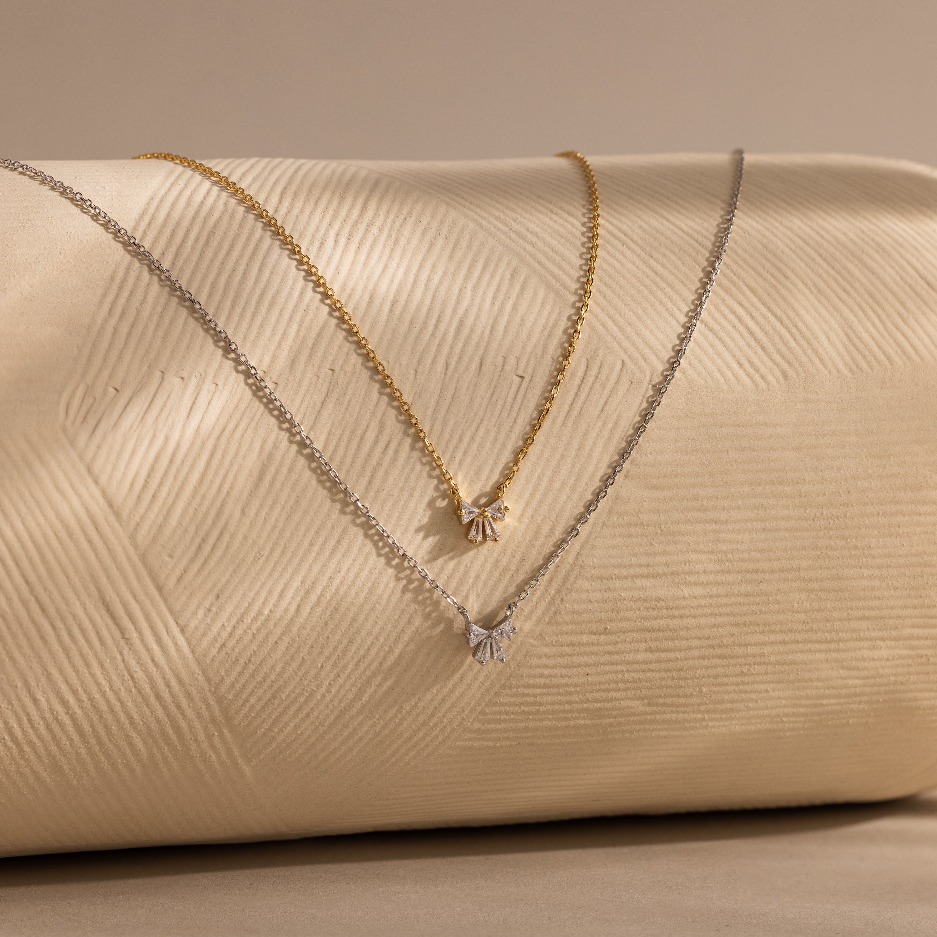 A delicate Diamond Bow Necklace in 18K Gold with a small bow-shaped pendant, displayed on a beige surface.