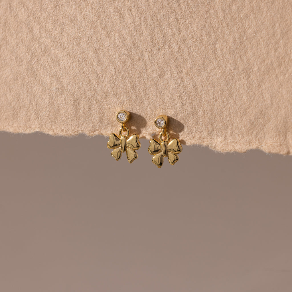 Bow Drop Studs in 18K Gold, featuring small round accents, are showcased on a textured beige surface.