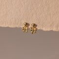 Bow Drop Studs in 18K Gold, featuring small round accents, are showcased on a textured beige surface.