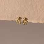 Bow Drop Studs in 18K Gold, featuring small round accents, are showcased on a textured beige surface.