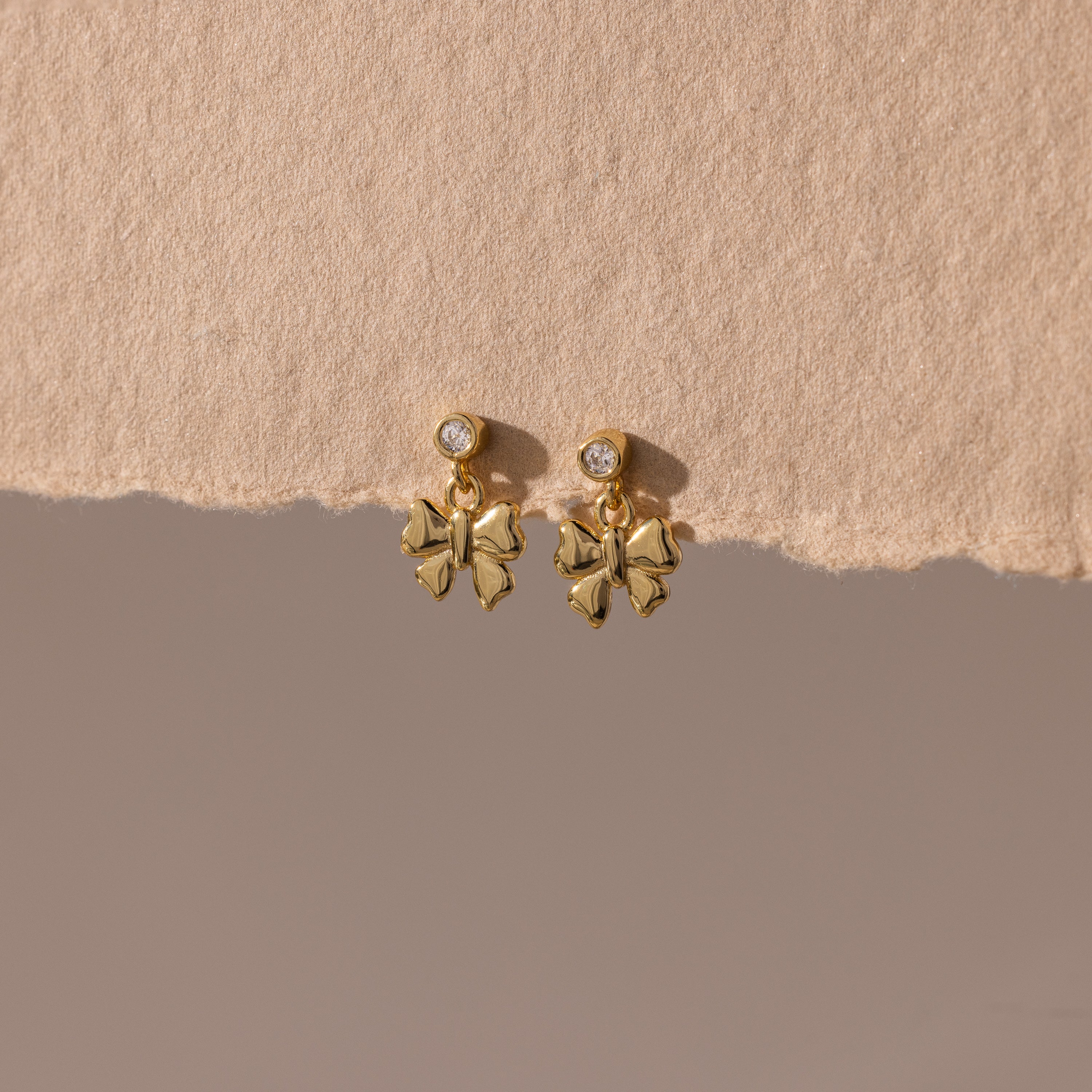 Bow Drop Studs in 18K Gold, featuring small round accents, are showcased on a textured beige surface.