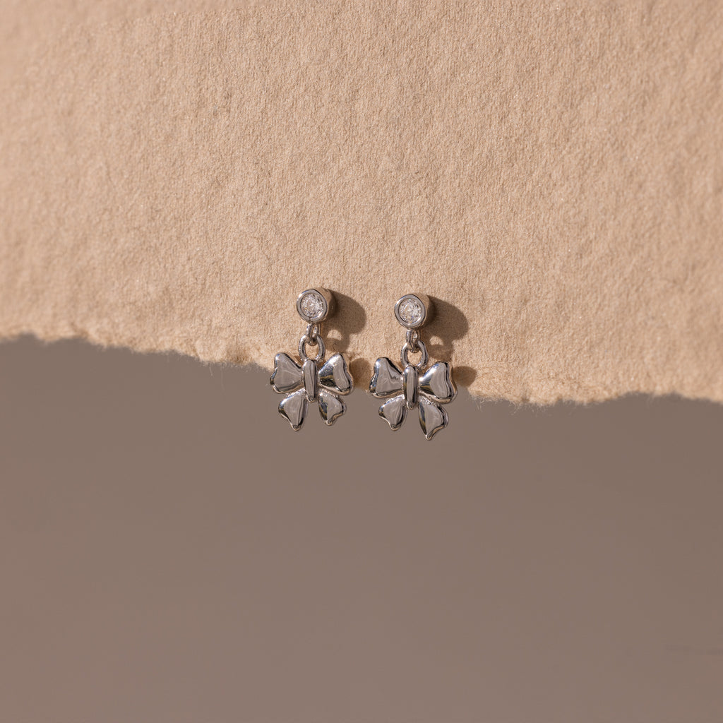 Bow Drop Studs in Sterling Silver, featuring small round accents, are showcased on a textured beige surface.