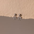 Bow Drop Studs in Sterling Silver, featuring small round accents, are showcased on a textured beige surface.