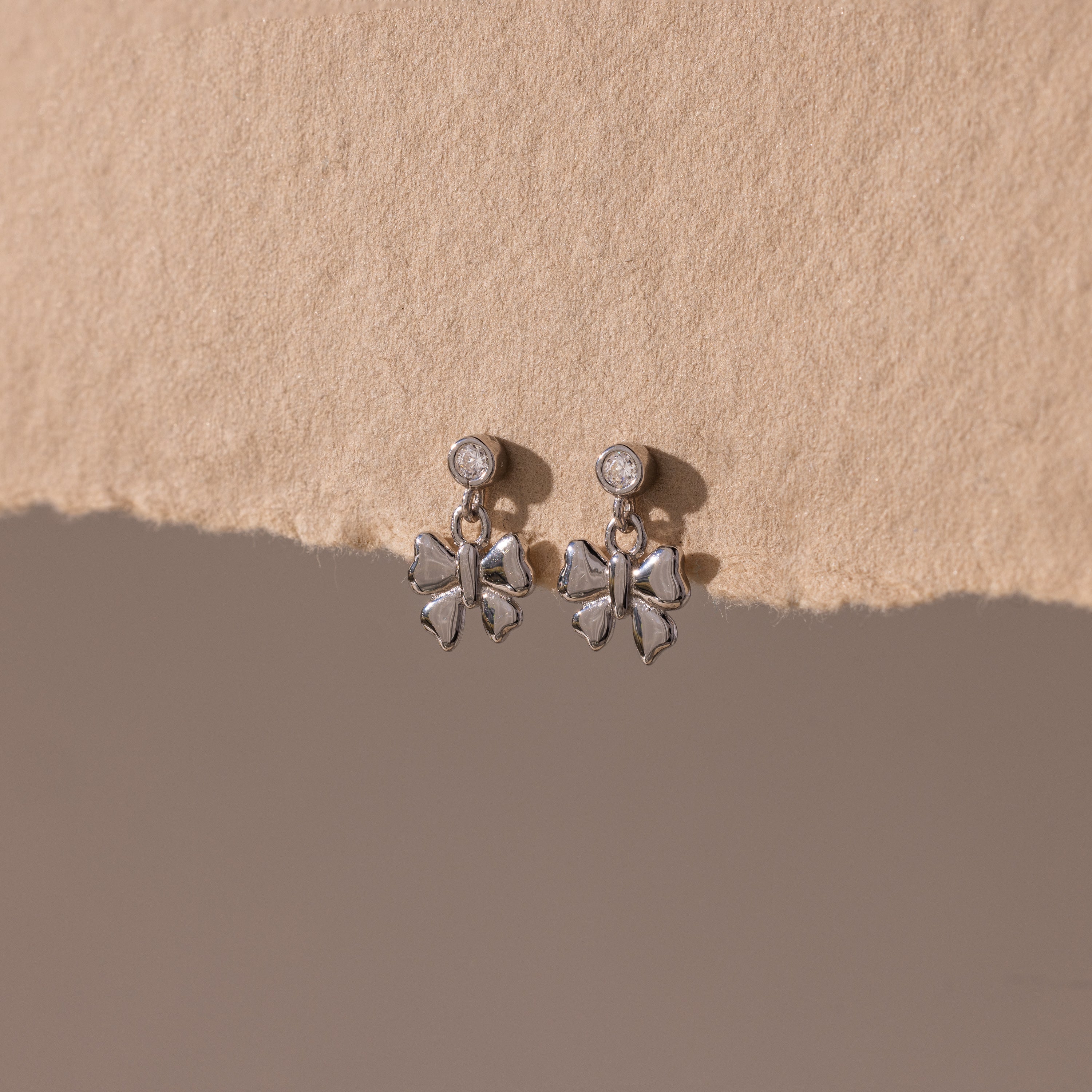 Bow Drop Studs in Sterling Silver, featuring small round accents, are showcased on a textured beige surface.