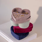 Stack of velvet heart-shaped jewelry boxes in lavender, mint green, red, and blue, with the top box displaying gold rings, necklace, and bracelets.