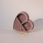 Lavender velvet heart-shaped jewelry box displayed upright, holding gold rings and a heart pendant necklace, with gold bangles placed in front.