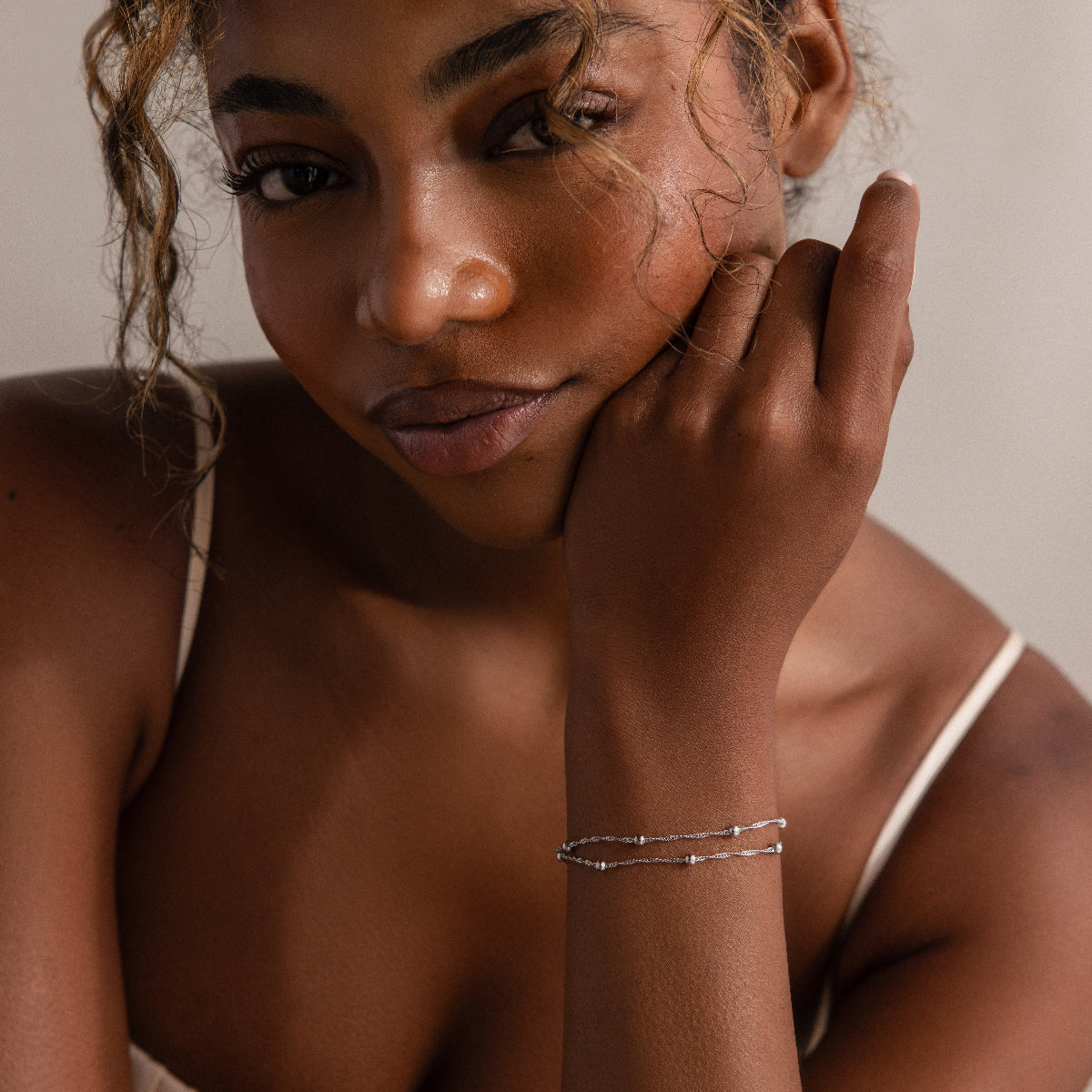 Woman with a hand on her face wearing a silver beaded bracelet against a neutral background