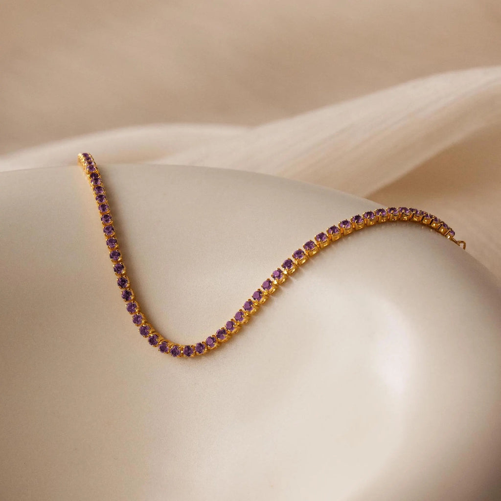 The Classic Birthstone Tennis Bracelet, a gold chain adorned with small purple birthstones, is elegantly draped over a smooth, cream-colored surface for a luxurious look.