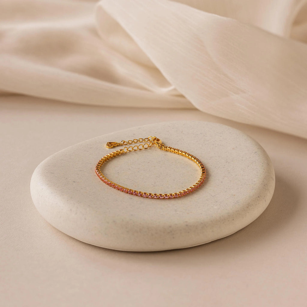 The Classic Birthstone Tennis Bracelet, adorned with pink gemstones, exudes delicate luxury and is elegantly displayed on a round stone over a beige fabric background.