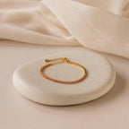 The Classic Birthstone Tennis Bracelet, adorned with pink gemstones, exudes delicate luxury and is elegantly displayed on a round stone over a beige fabric background.