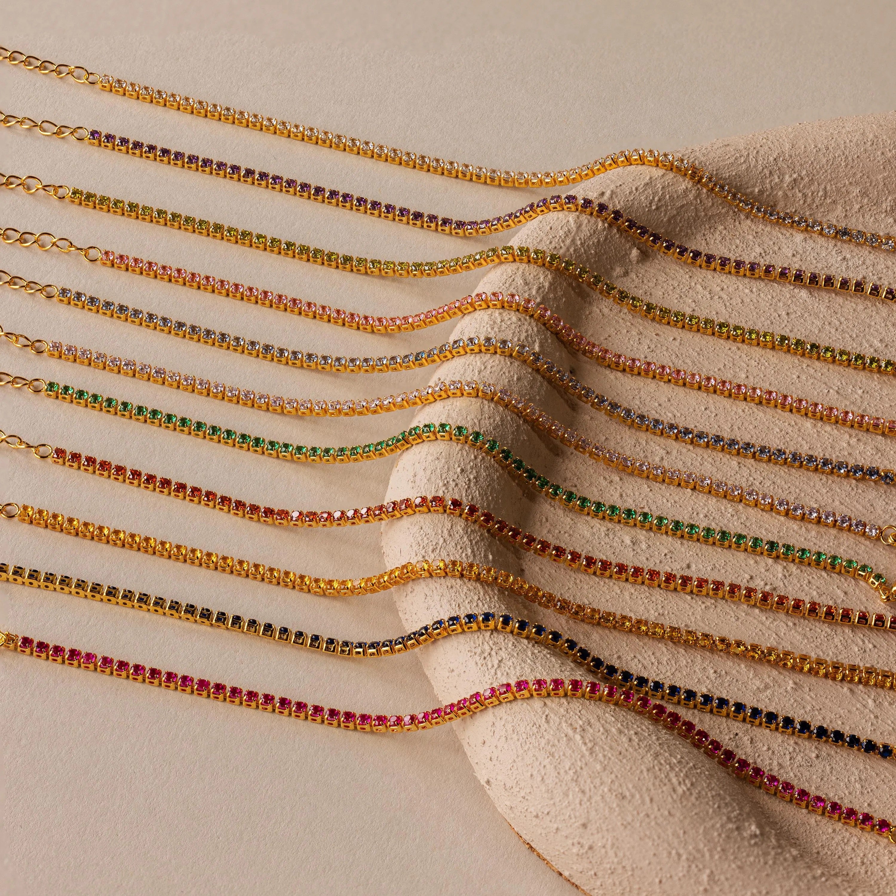 The Classic Birthstone Tennis Bracelet is arranged in a parallel line with other gold chain gemstone bracelets on a textured beige surface.