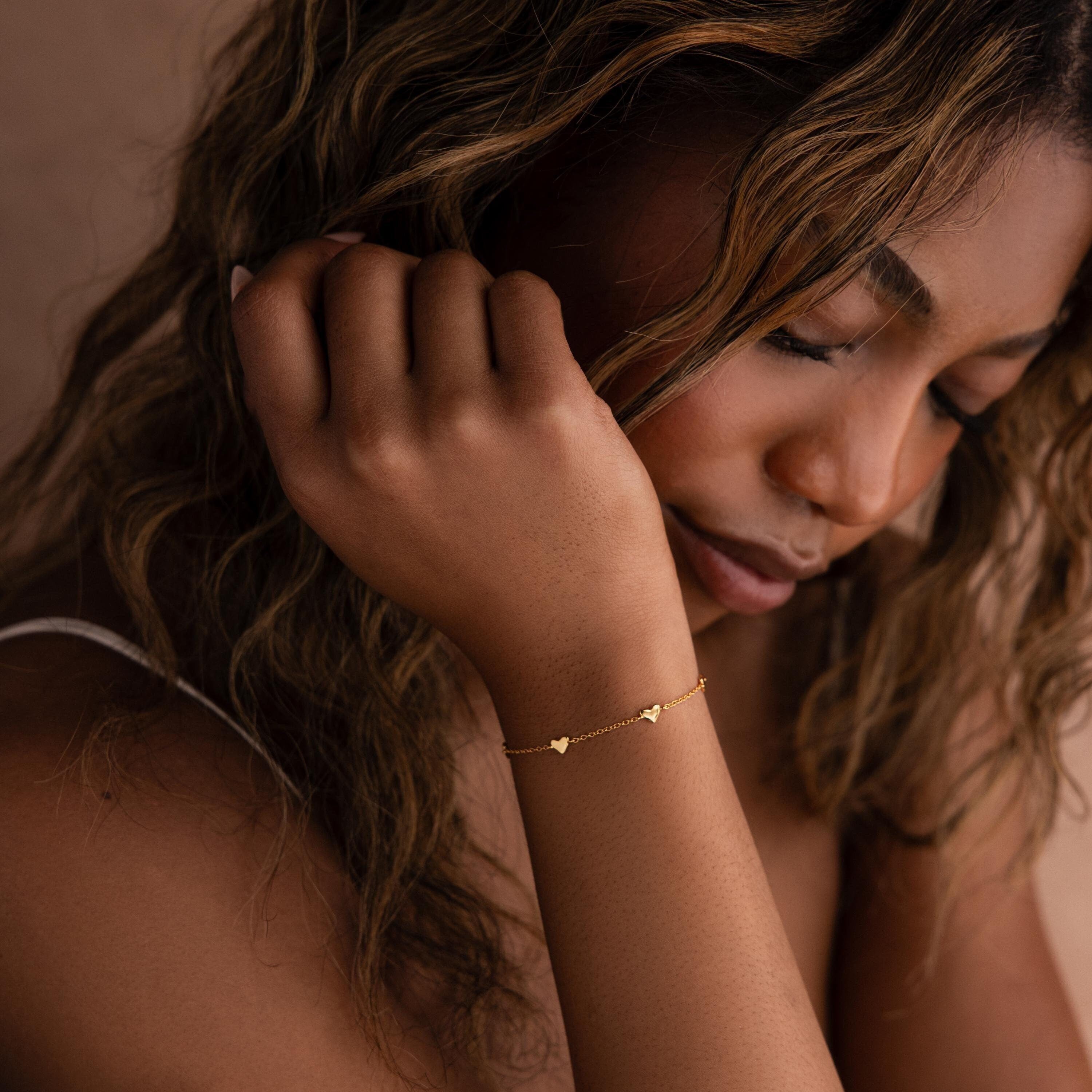 A woman with wavy hair gazes downward, wearing the Heart Station Bracelet. The delicate piece adds a romantic and graceful touch to her style.