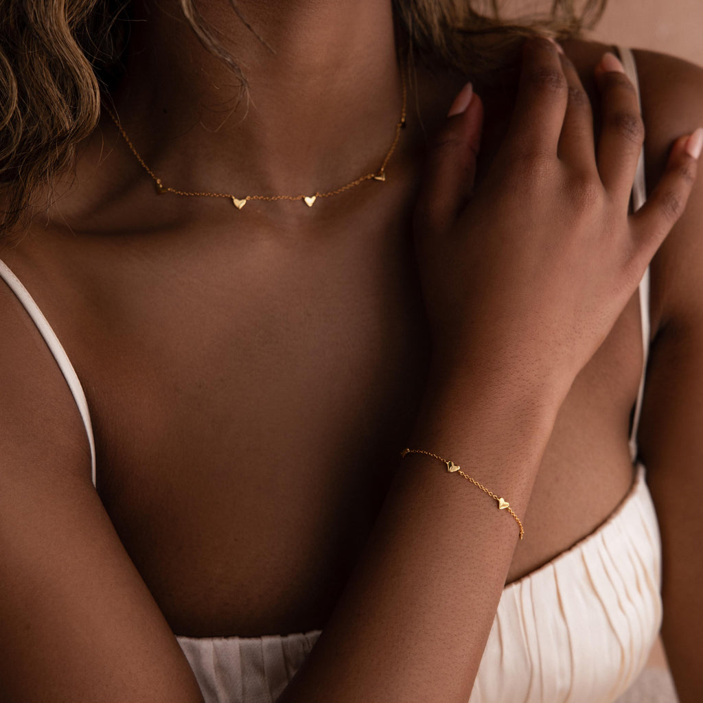 A woman in a white top touches her shoulder gently while wearing a gold heart necklace and the elegant Heart Station Bracelet.