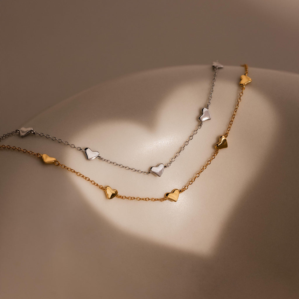 The Heart Station Bracelet, available in gold and silver, is stackable for a chic layered look. Displayed on a smooth surface with soft lighting, it's the perfect romantic accessory for any occasion.
