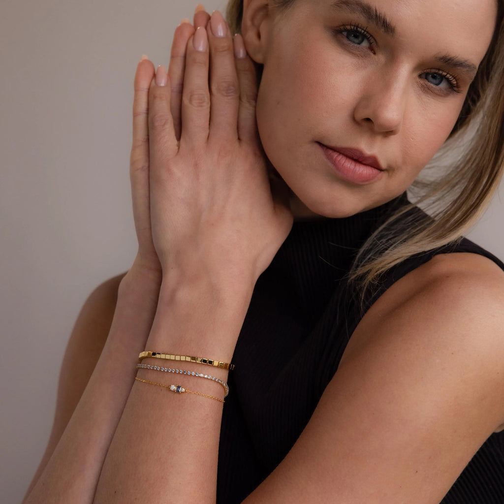 A woman in a sleeveless black top clasps her hands, highlighting gold and silver bracelets on her wrist, including the striking Cube Bracelet featuring geometric bead links.