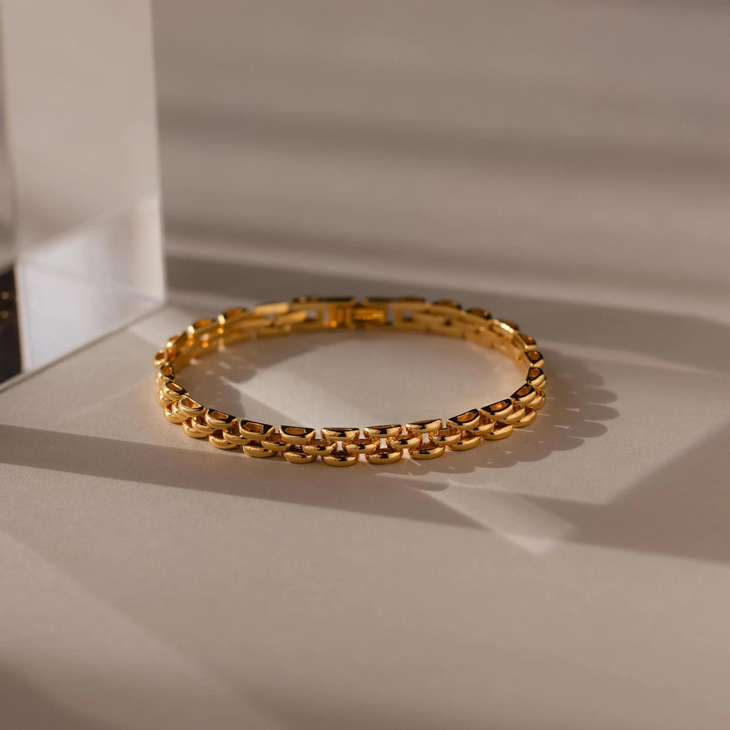 A Strap Bracelet with a textured link design rests on a beige surface in soft, shadowed lighting, highlighting it as a bold statement piece.