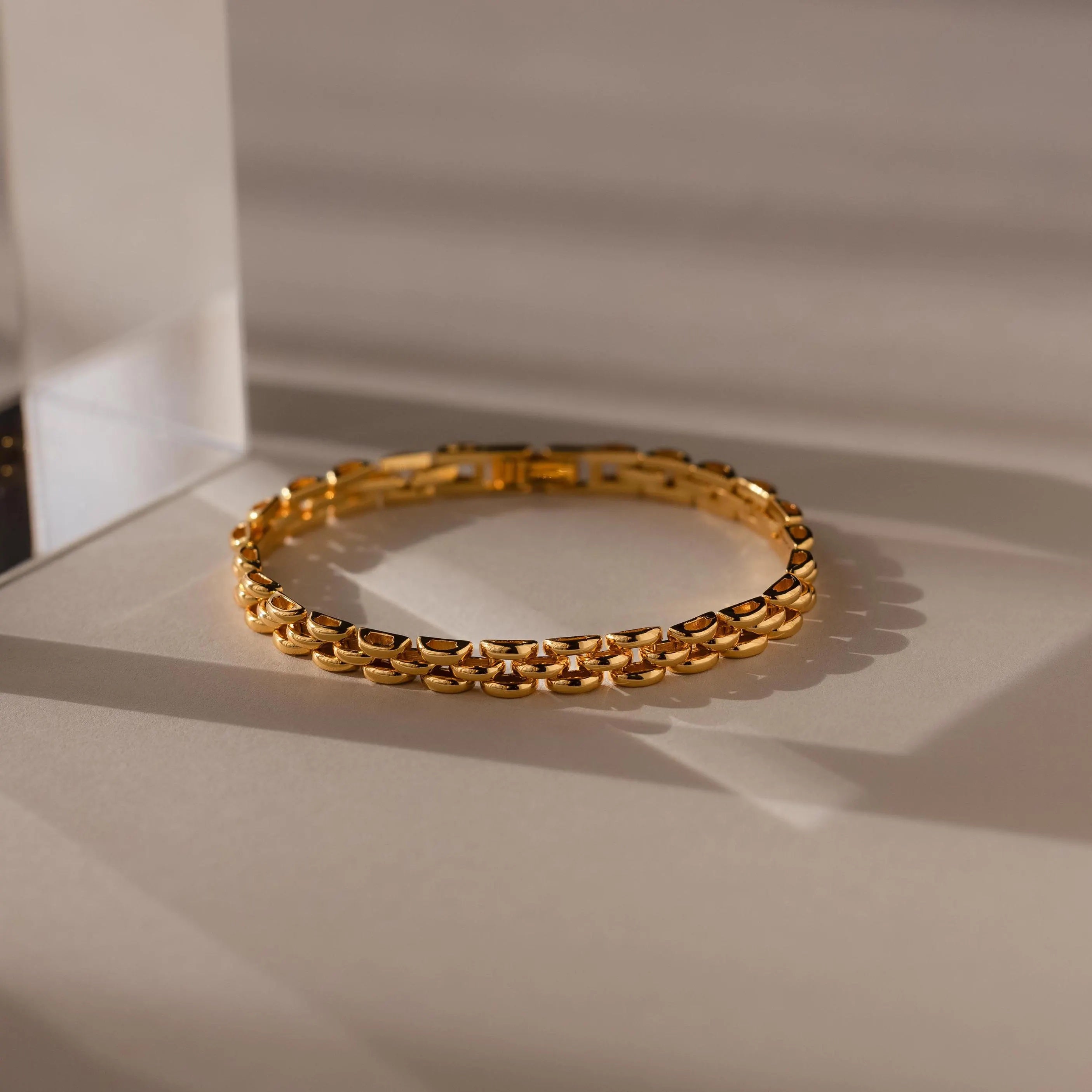 A Strap Bracelet with a textured link design rests on a beige surface in soft, shadowed lighting, highlighting it as a bold statement piece.
