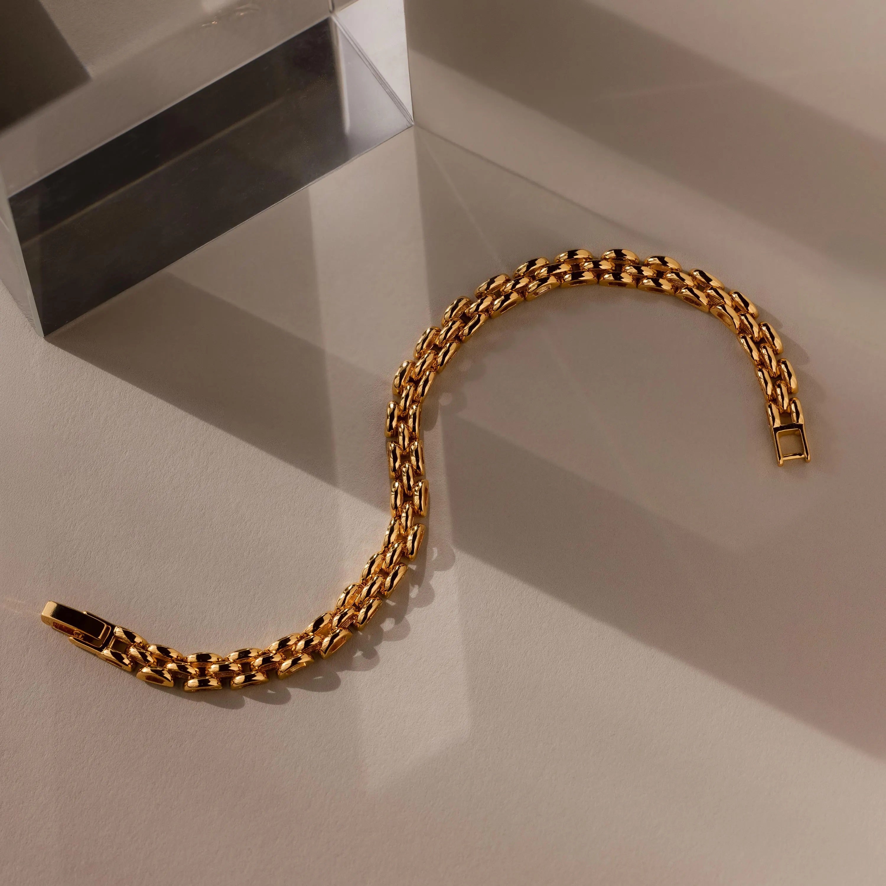 A Strap Bracelet with a gold panther chain design lies in an S shape on a beige surface beside a clear rectangular block, creating a striking statement piece.