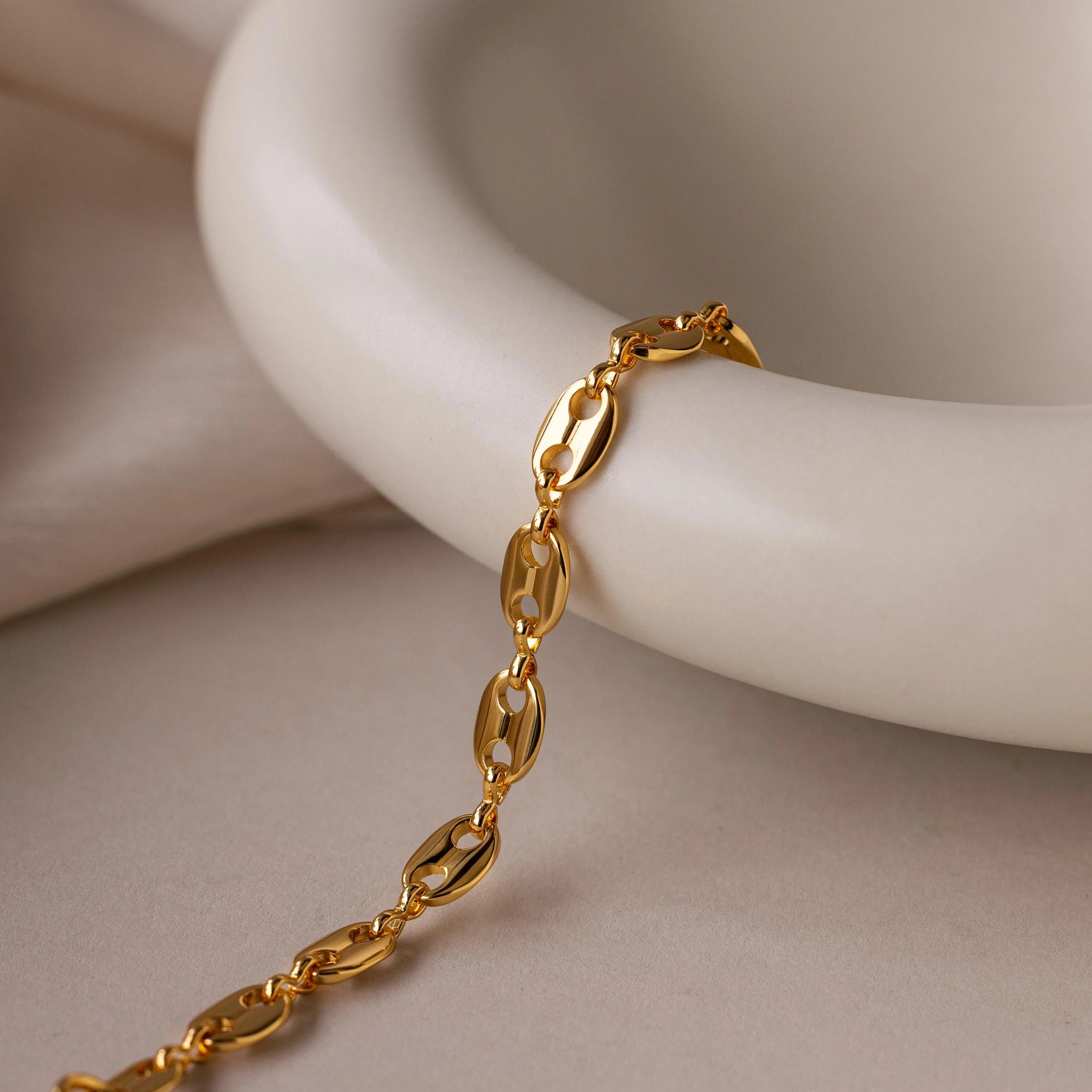 The Bold Mariner Chain Bracelet, featuring oversized links, drapes elegantly over a smooth cream ceramic bowl—an eye-catching statement jewelry piece.