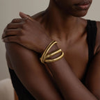 Wearing three chunky gold bracelets, including the Ribbed Infinity Bangle, a person crosses their arm over their chest to make a chic style statement.