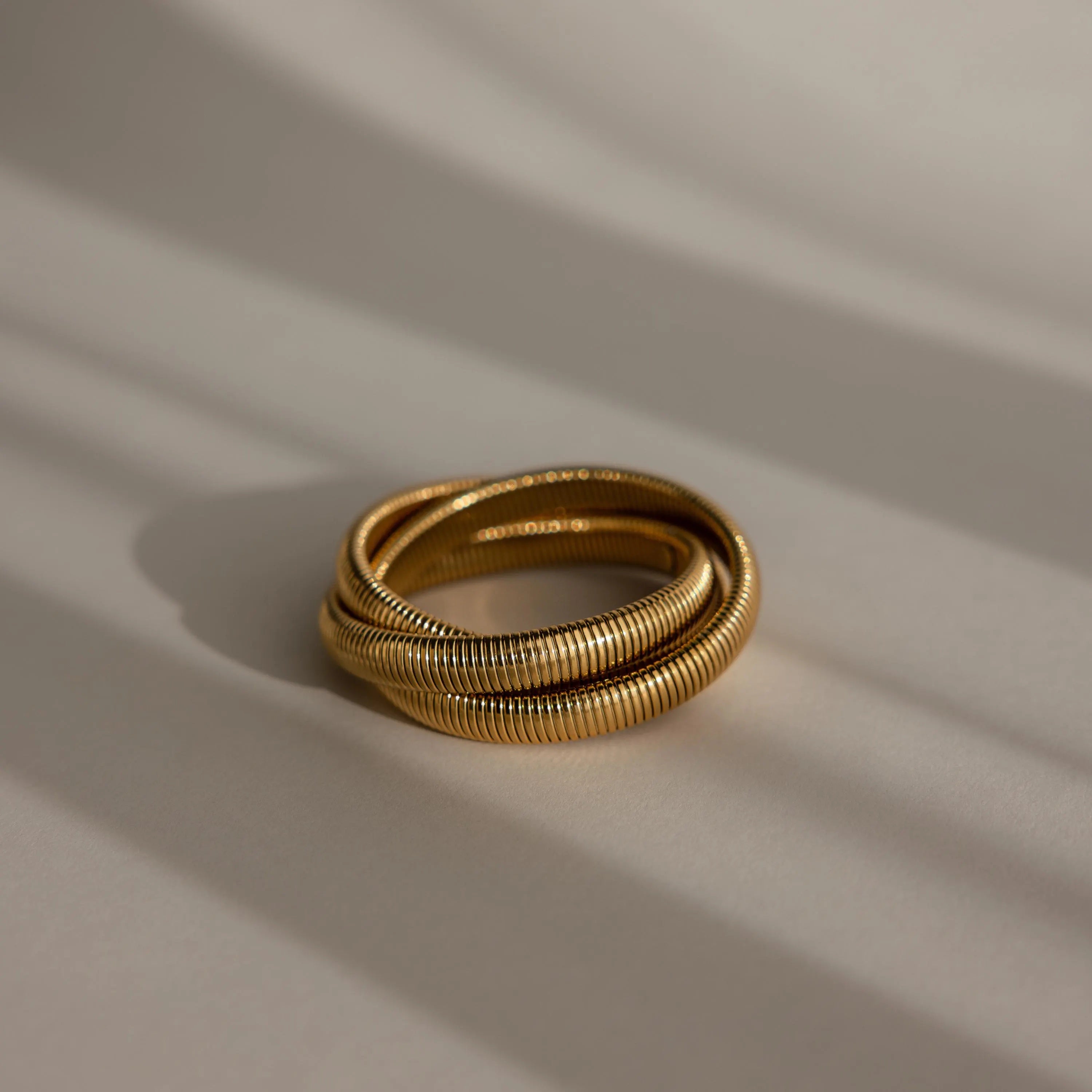 The Ribbed Infinity Bangle, featuring three interlocking gold rings with a ribbed texture, is displayed on a soft beige background with gentle shadows.