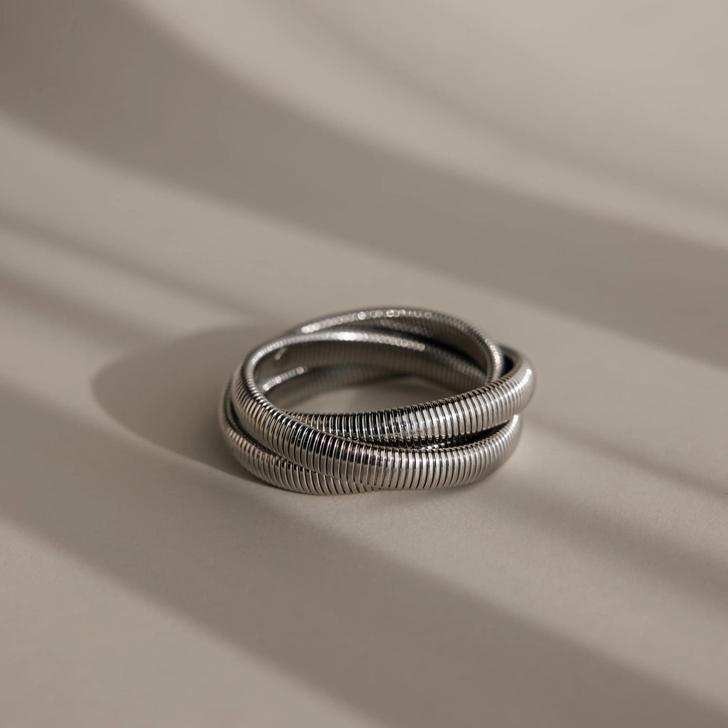 The Ribbed Infinity Bangle is a coiled silver bracelet with a ribbed texture, elegantly displayed on a softly shadowed, neutral surface.