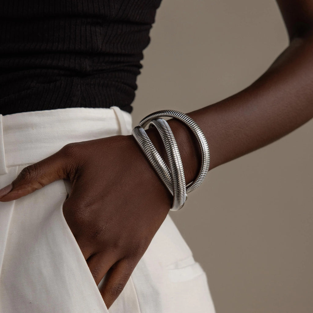 Dressed in white pants and a black top, someone sports chunky silver bracelets on their wrist, featuring the Ribbed Infinity Bangle for a stylish look.