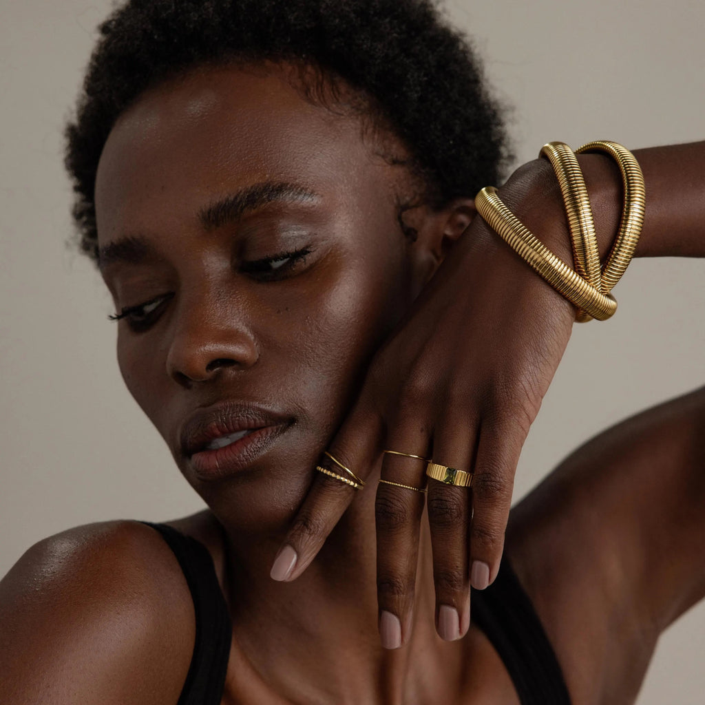 A woman with dark skin poses against a plain background, wearing stylish rings and gold bracelets, including the Ribbed Infinity Bangle, as she holds her hand near her face.