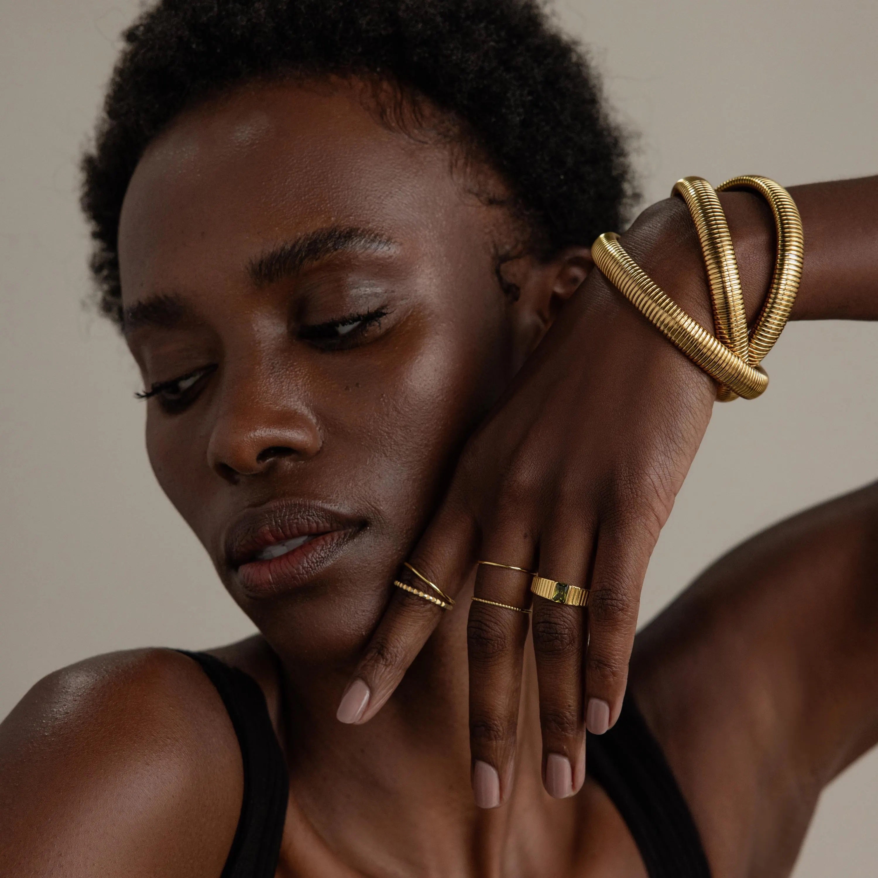 A woman with dark skin poses against a plain background, wearing stylish rings and gold bracelets, including the Ribbed Infinity Bangle, as she holds her hand near her face.