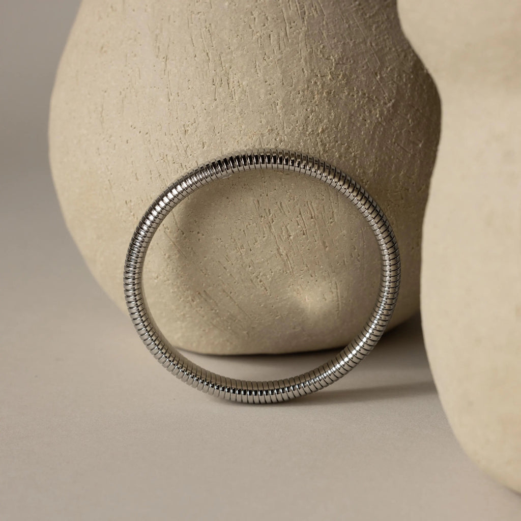 The Thick Snake Chain Bangle in silver is displayed beside two textured light beige ceramic vases on a neutral background.