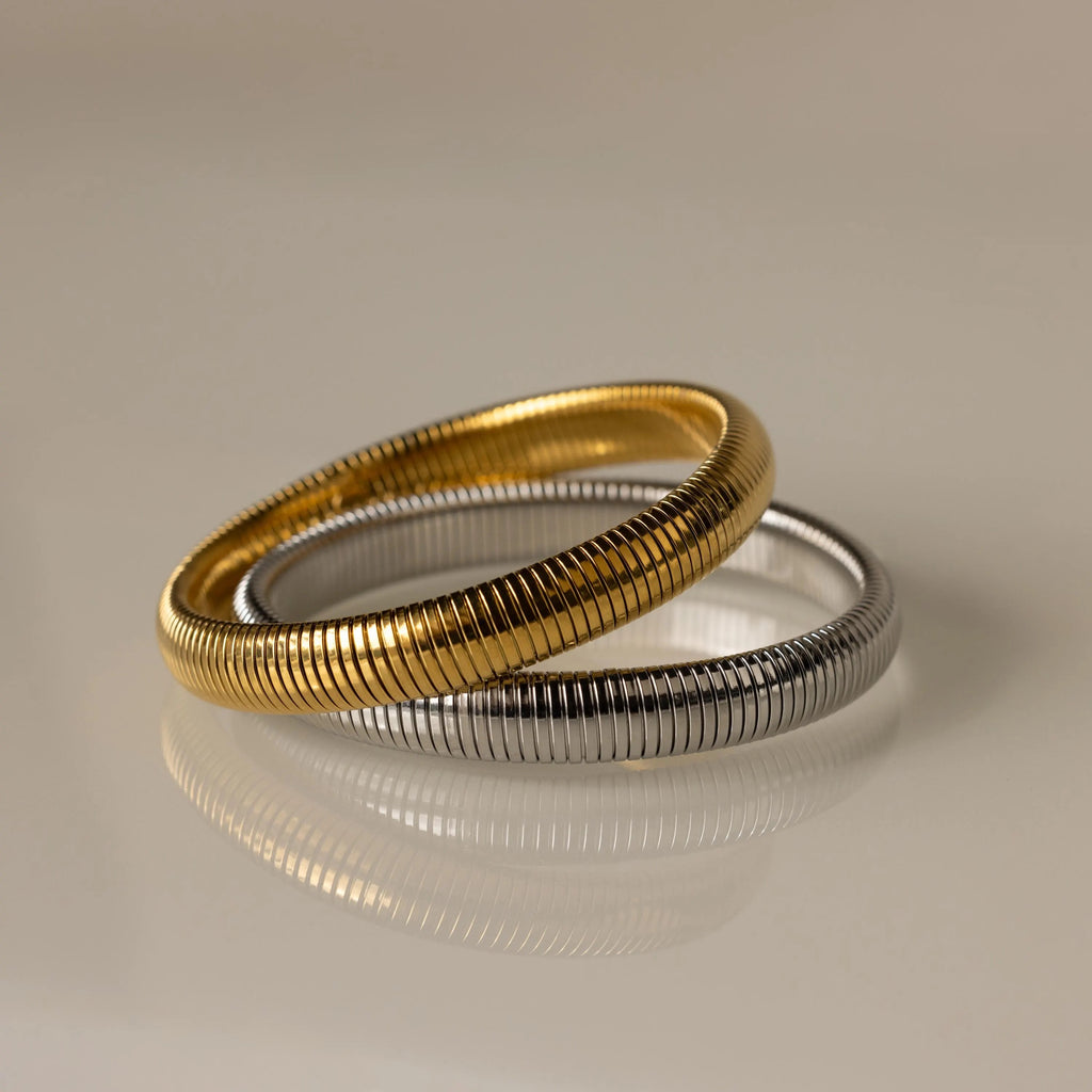 Two Thick Snake Chain Bangles, one gold and one silver, with a textured design overlap on a smooth, reflective beige surface.