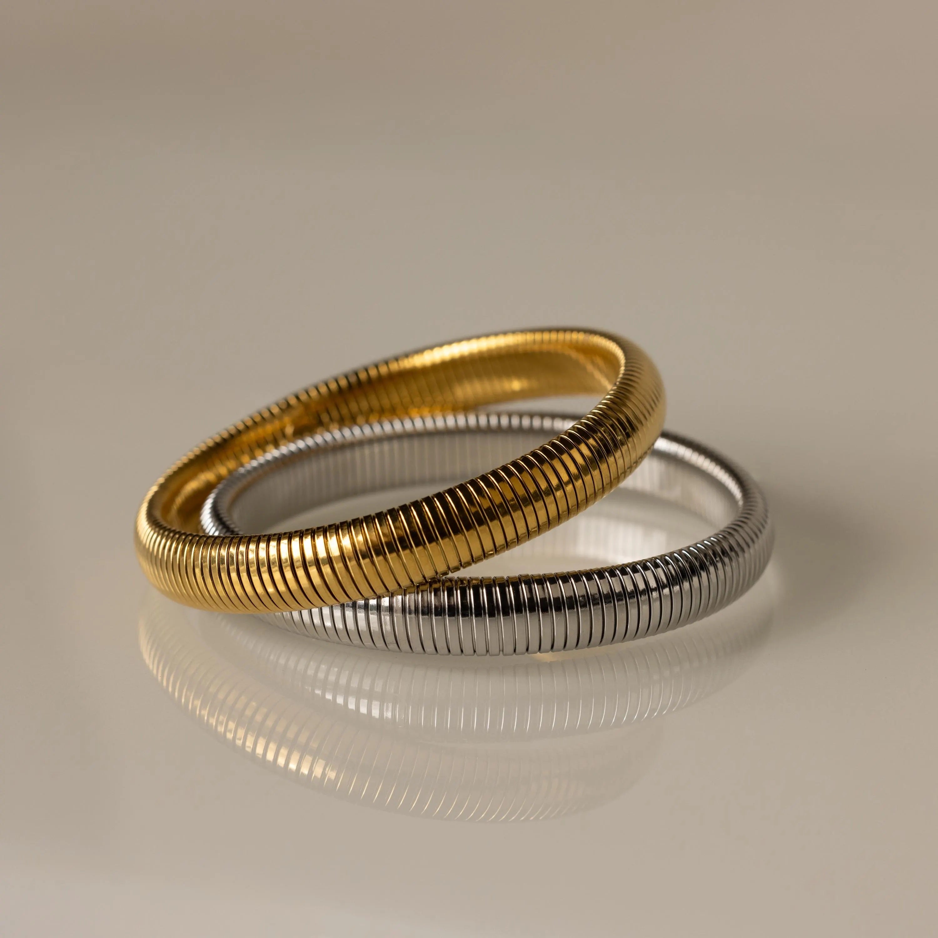 Two Thick Snake Chain Bangles, one gold and one silver, with a textured design overlap on a smooth, reflective beige surface.