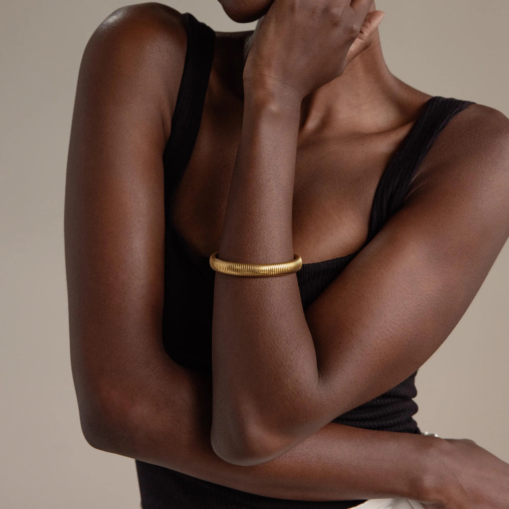 A woman in a black tank top wears the Thick Snake Chain Bangle, featuring a ribbed pattern, with her arm crossed over her chest against a neutral background.