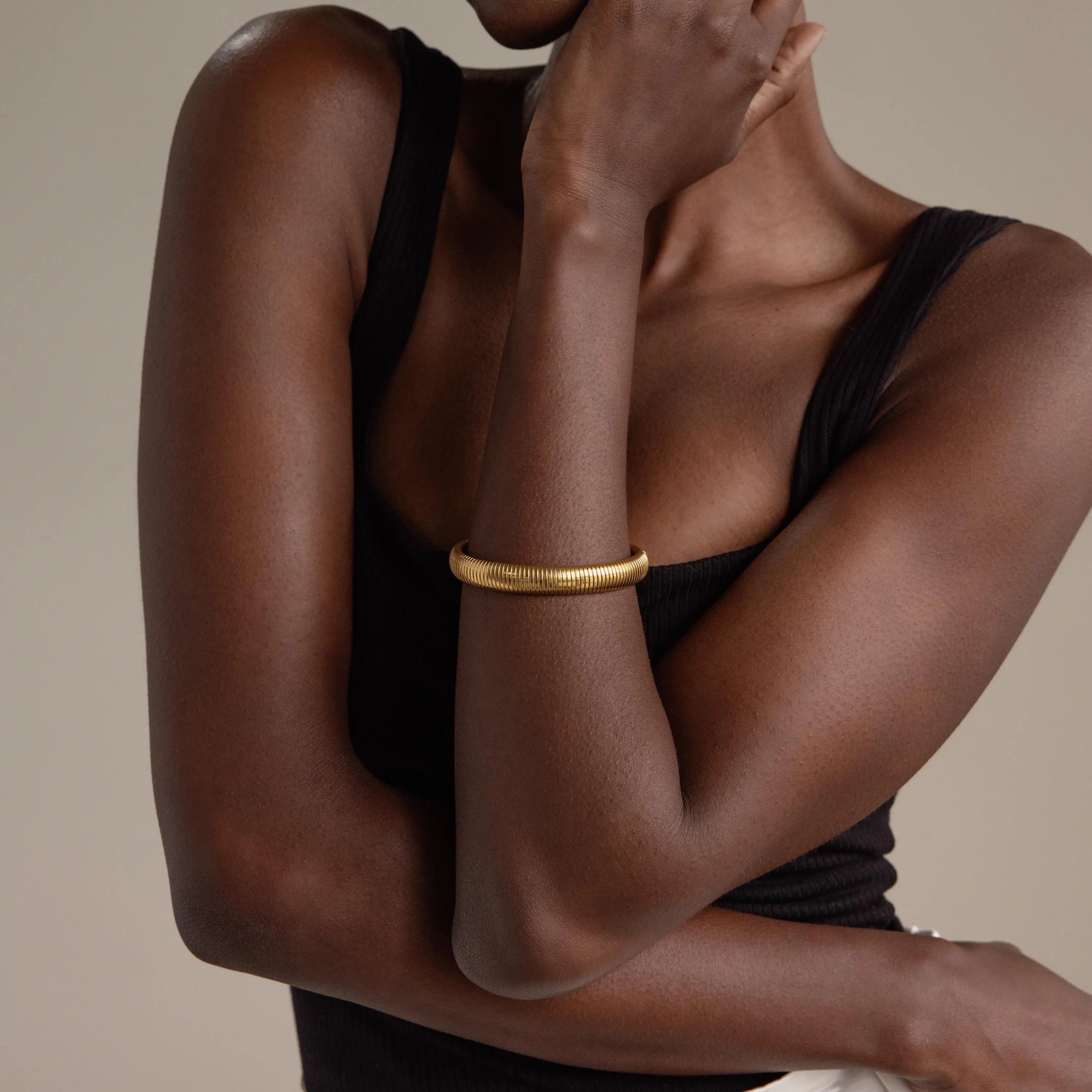 A woman in a black tank top wears the Thick Snake Chain Bangle, featuring a ribbed pattern, with her arm crossed over her chest against a neutral background.